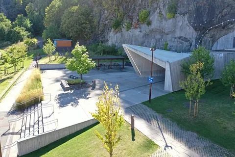 Moderne bygg ved fjellvegg med grøntanlegg, sittegrupper og gangstier i solfylt parkmiljø.